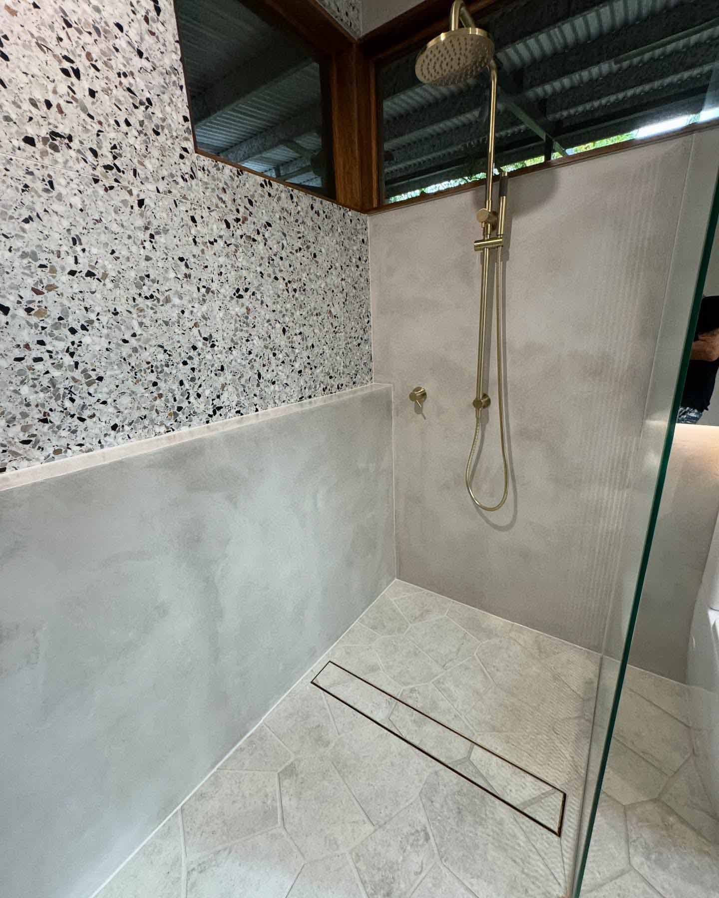 Walk-in shower with smooth grey walls, a terrazzo-patterned upper wall and brass fittings